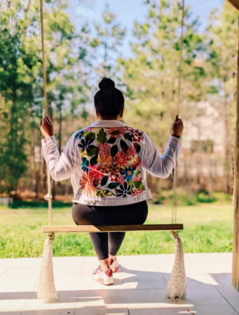 Woman wearing a Tabitha Sewer floral denim jacket sitting on a swing, with her back to the camera.