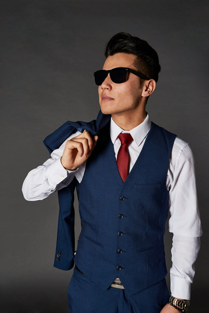 Man wearing a five-button blue waistcoat over a white dress shirt and red tie, with suit jacket draped over his right shoulder, and wearing dark sunglasses. 